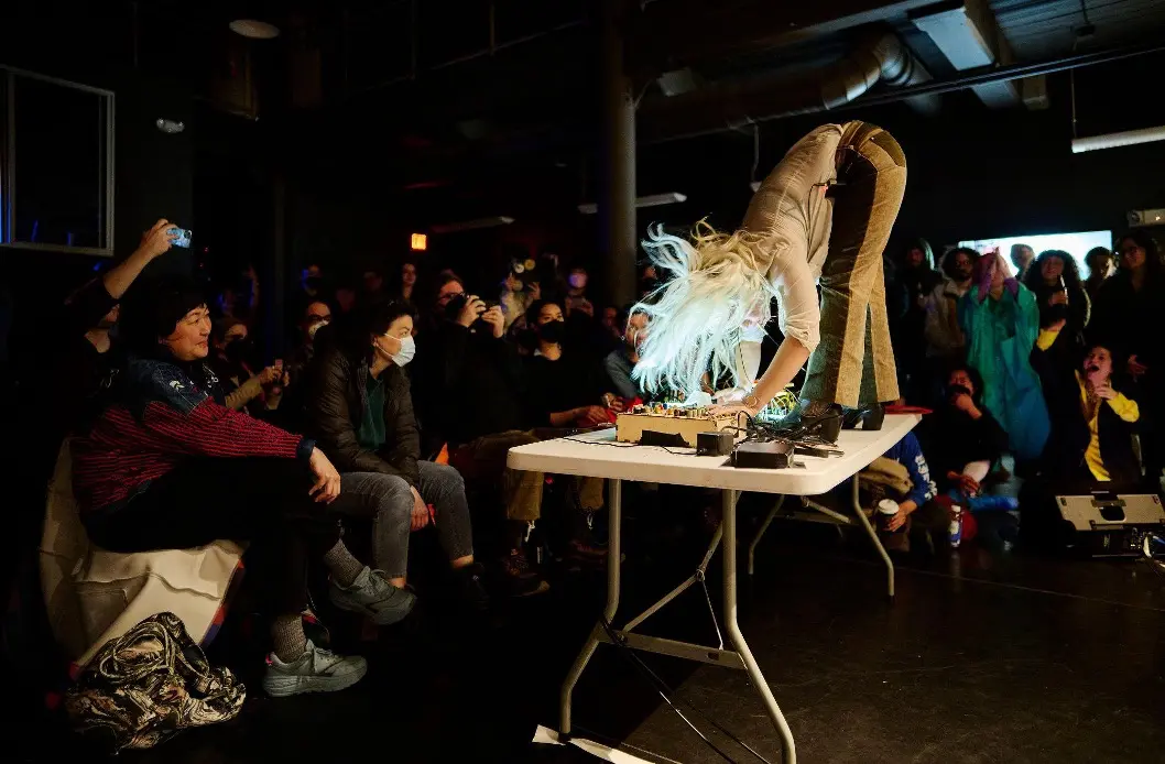DJ Victoria Shen stands on a fold-out table, bent over at the waist to reach her controller while a crowd looks on