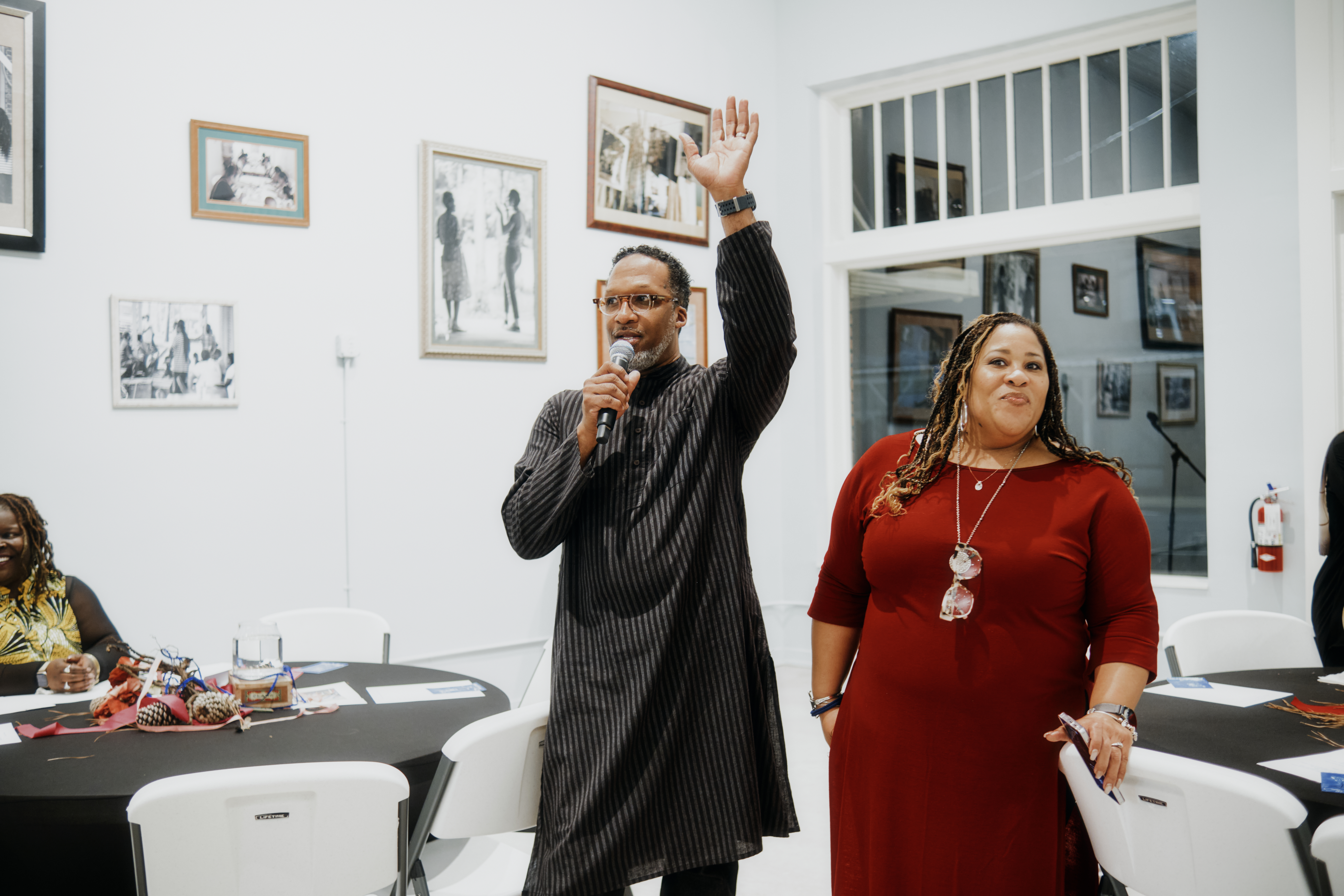 Carlton, on the left, is wearing a gray kaftan and speaking into a microphone with his hand raised. Brandi is standing beside him in a red dress, with her hand on a chair, smiling.