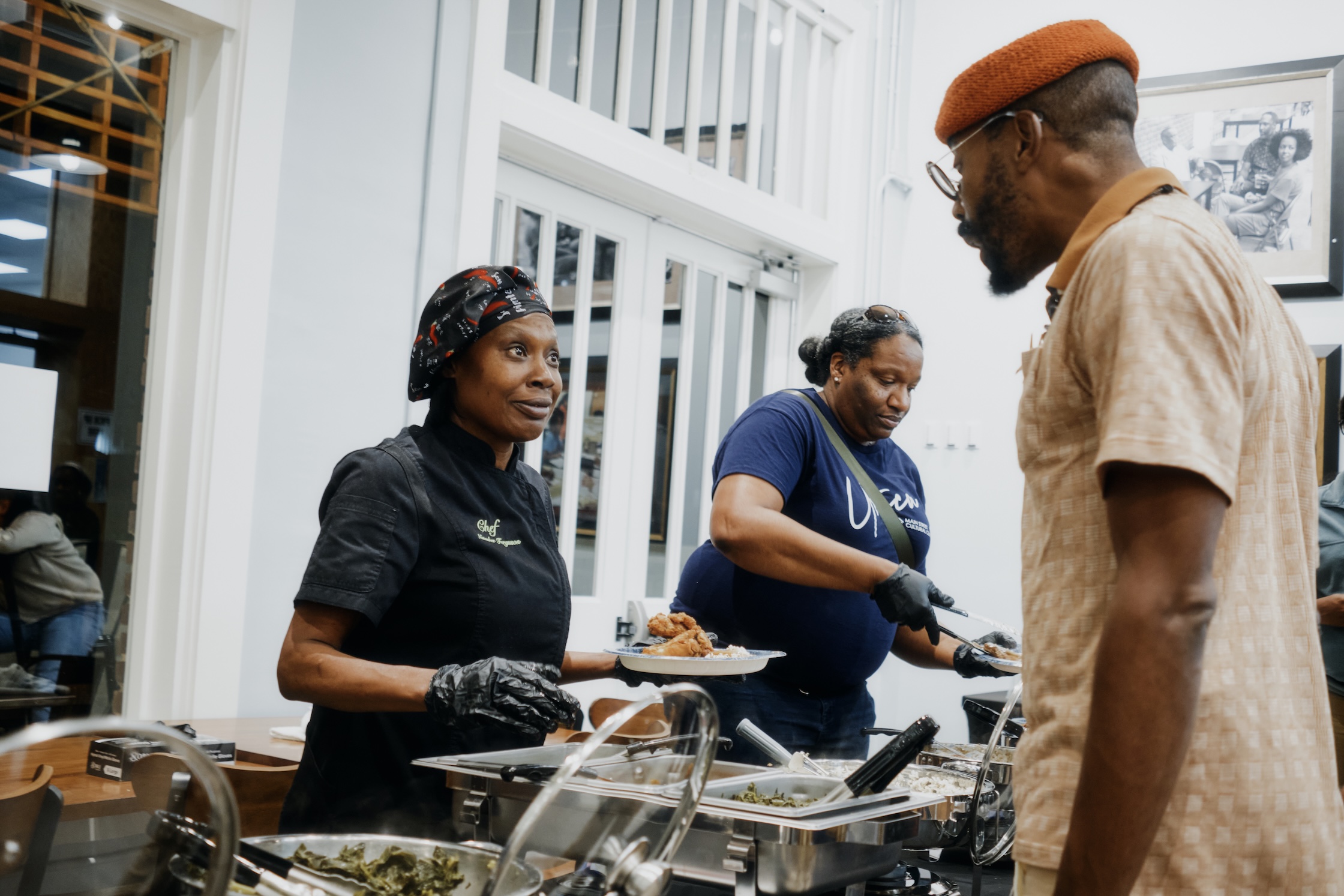 Natasha is in the foreground of the photo, handing food to a man with an umber beret on, who is talking to her across the food serving line.