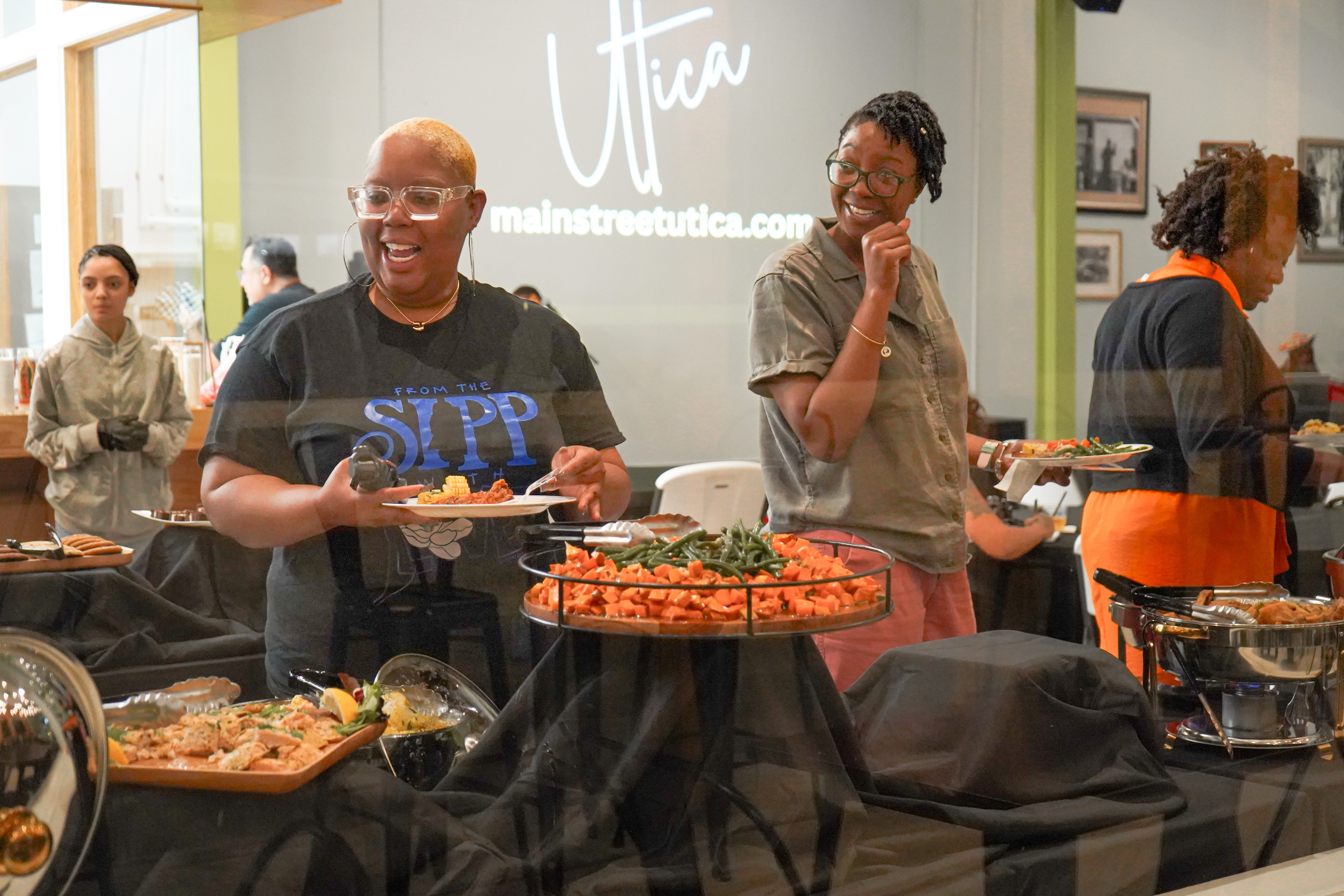 Jasmine is in the center of the photo, reaching out toward a tray of food and smiling. She has large framed glasses and short white-blonde hair, and her shirt says SIPP.